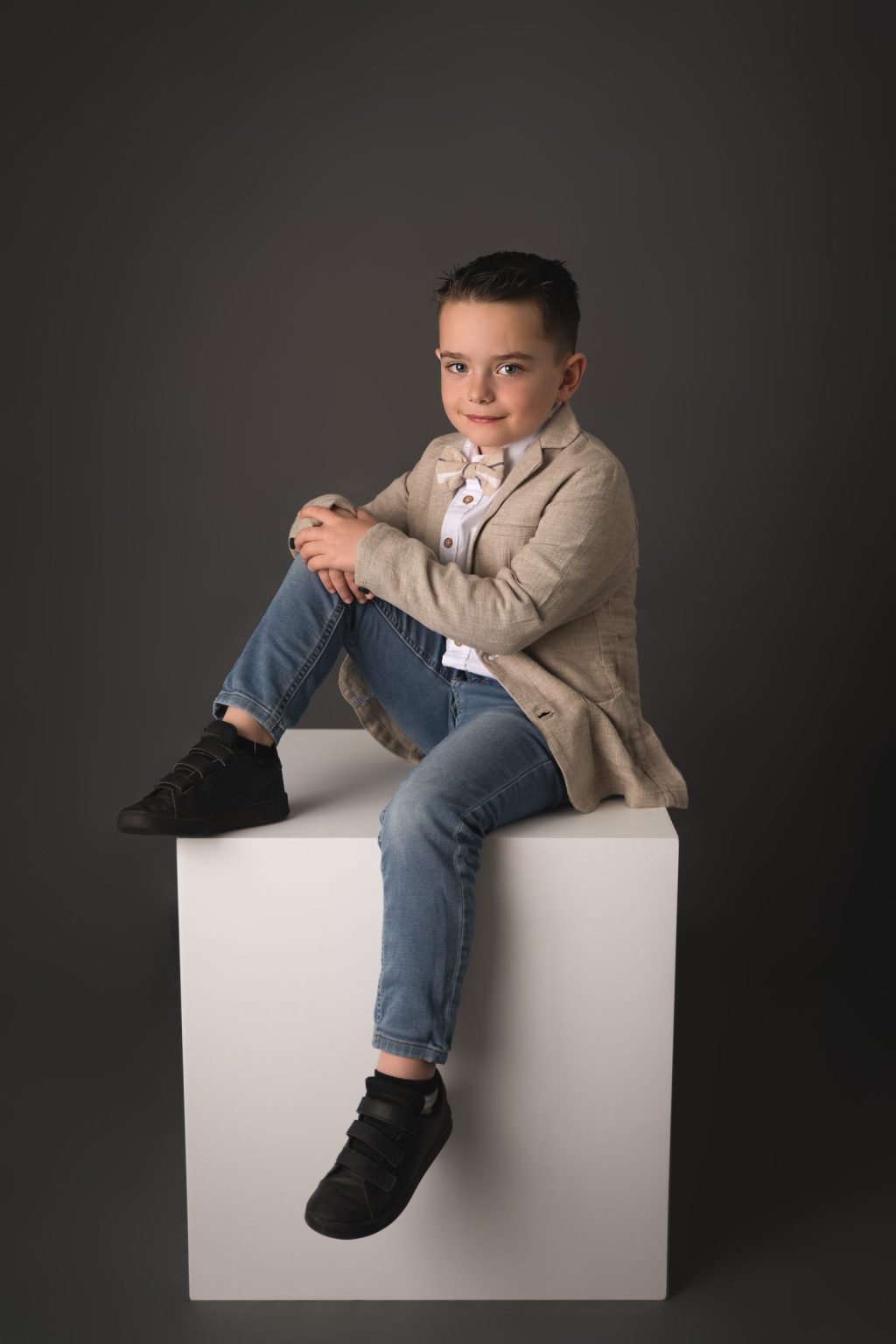 Petit gar&ccedil;on en veste beige assis sur un cube blanc, fond gris &ndash; s&eacute;ance photo enfant &agrave; Tournai
